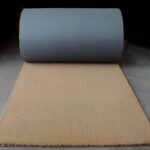 Rullo di tappeto in fibra naturale su pavimento grigio, pronto per essere steso e utilizzato in interni al cocco naturale di colore ocra