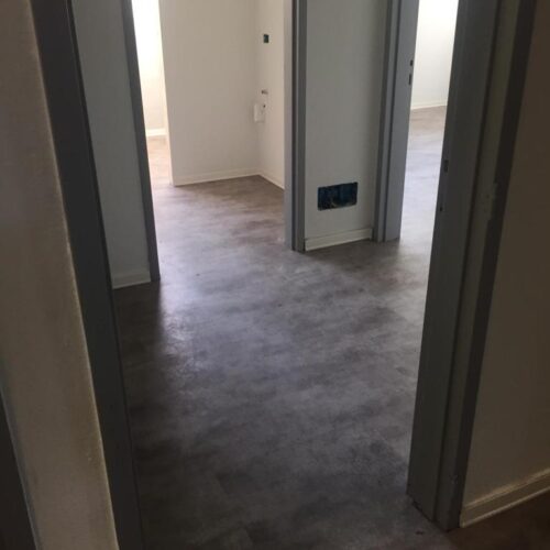 Corridoio di una casa con pavimento grigio in cemento, porte aperte che conducono a stanze luminose.