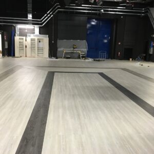 Un'ampia sala interna con pavimento laminato chiaro e strisce scure, pareti nere e attrezzature tecniche sullo sfondo.