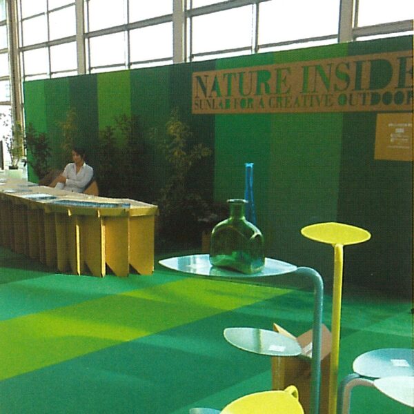 Stand espositivo con tema ecologico, decorato con piante verdi e tavoli, uomo seduto al banco. Natura dentro.