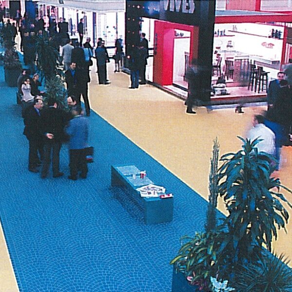 Un'area espositiva coperta con stand commerciali, persone che conversano e piante decorative su pavimentazione colorata.