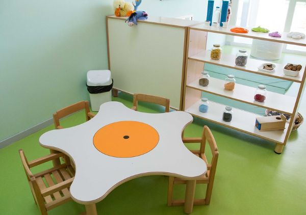 Una stanza per bambini con tavolo bianco e arancione, scaffali con giochi educativi e pavimento verde chiaro.