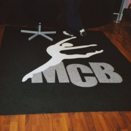 Tappeto nero con il logo MCB in bianco e la silhouette di una ballerina che salta, pavimento in parquet