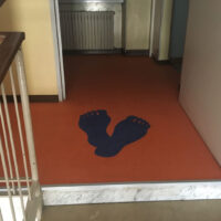 Immagine di un tappetino arancione con impronte blu a forma di piedi in un corridoio di un edificio.
