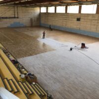 Grande palestra al chiuso in fase di ristrutturazione con operai che lavorano al rifacimento del pavimento in legno.