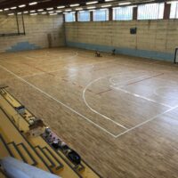 Palestra vuota con pavimento in legno lucido, tribune gialle e pareti beige. Luce naturale dalle finestre.