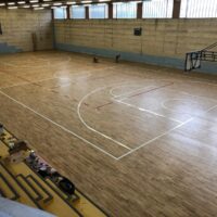 Campo da basket al coperto con pavimento in legno, gradinate vuote e alcune attrezzature sportive a bordo campo.