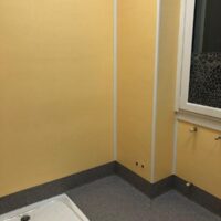 Stanza vuota con pareti gialle, pavimento grigio e una piccola finestra. Nell'angolo c'è una cabina doccia.