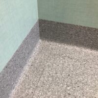 Angolo di una stanza con pareti verde chiaro e pavimento in linoleum grigio puntinato che si estende sui muri.