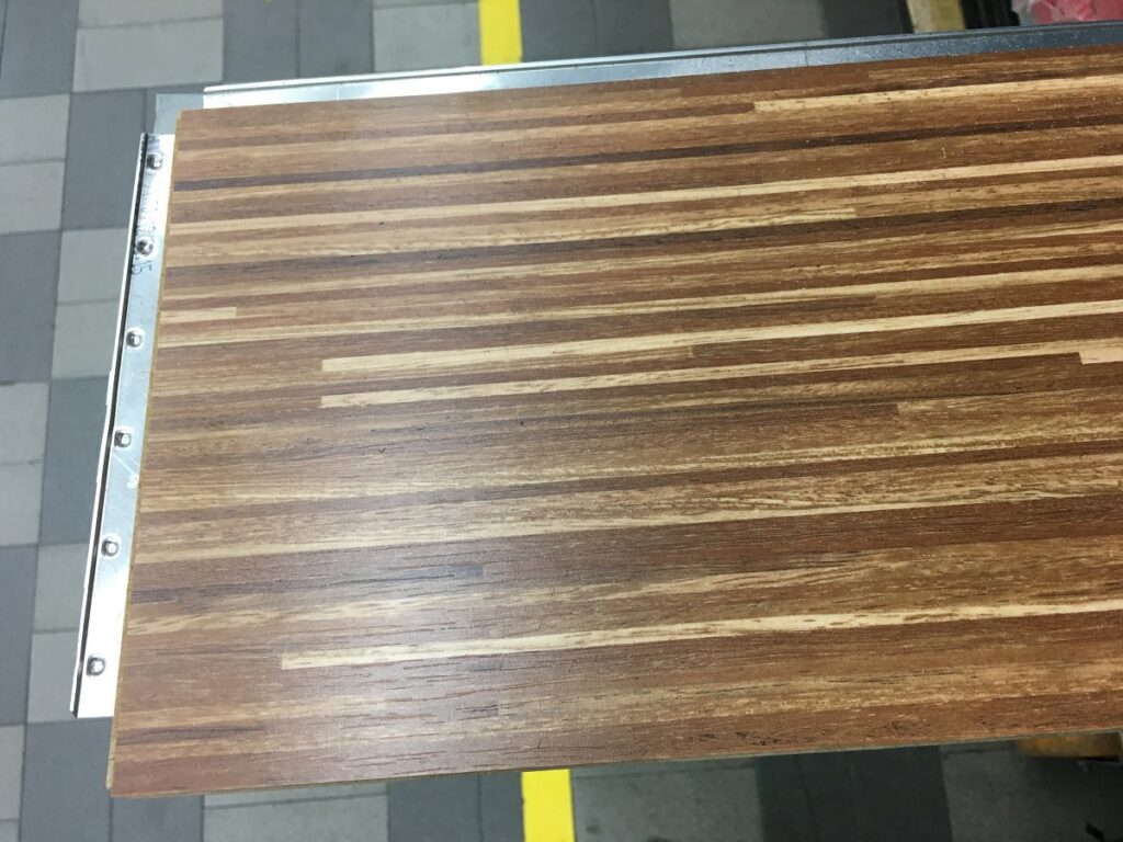 Tavolo di legno con superficie a strisce orizzontali di varie tonalità marrone, posizionato su un pavimento a quadri.
