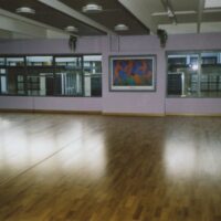 Sala spaziosa per danza con pavimento in legno chiaro, pareti lilla e specchi grandi che riflettono la luce.