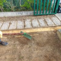Persona in abiti da lavoro sistemando il terreno con una vanga vicino a una recinzione verde in un giardino.