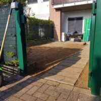 Cortile di una casa con pavimentazione in pietra e terra battuta, attrezzi da giardino e vegetazione sul lato.