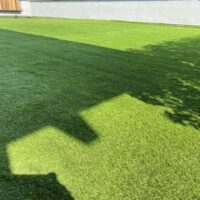 Prato verde curato con ombre e sole, adiacente a un muro chiaro e una struttura in legno visibile a sinistra.