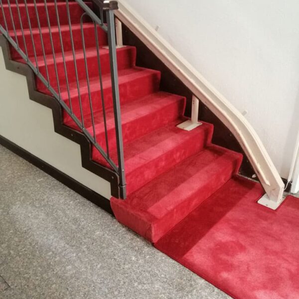 Scala rivestita di moquette rossa con corrimano metallico su un lato e pavimento in parquet grigio chiaro.